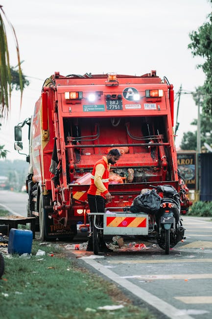A worker dressed in a red and yellow uniform is operating a large red rubbish collection truck on the side of a street. The truck's back panel is open, revealing the collection mechanism inside, which appears to be in the process of emptying or compacting waste. The worker is standing on a small platform, leaning slightly forward, with hands engaged near the waste compartment. Nearby, a black garbage bag rests on a small metal cart attached to a mobility scooter, positioned on the curb. The scene is set outdoors during daytime, with overcast weather, and the background includes trees, utility poles, and a few commercial buildings, indicating a suburban or urban area. Dirt and scattered rubbish are visible on the pavement around the truck, emphasizing the context of waste collection or waste management service. The overall scene aligns with private waste handling services often involved in rubbish removal and on-site clearance activities.