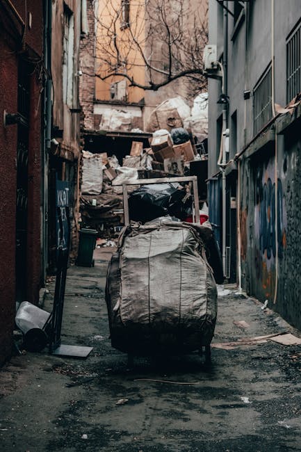 A narrow alleyway with a patchy, worn asphalt surface, cluttered with a large, grey, weathered fabric recycling or waste bag in the foreground, surrounded by small debris and discarded items. The alley is flanked by tall, gritty brick and concrete buildings with visible graffiti and signs of weathering on the walls, including pipes and electrical cables on the right side. At the end of the alley, there is an accumulated pile of various waste materials, including cardboard boxes, plastic bags, and other refuse, forming a makeshift informal rubbish dump. The environment appears urban and unmanaged, suggesting it may be an area where private waste disposal services are employed by Waste Disposal Catford or similar companies for on-site clearance, rather than standard municipal collection. Sparse, leafless street trees are visible in the background, with a diffused natural light creating a somewhat bleak atmosphere typical of such alleyways used for waste disposal and rubbish removal practices.