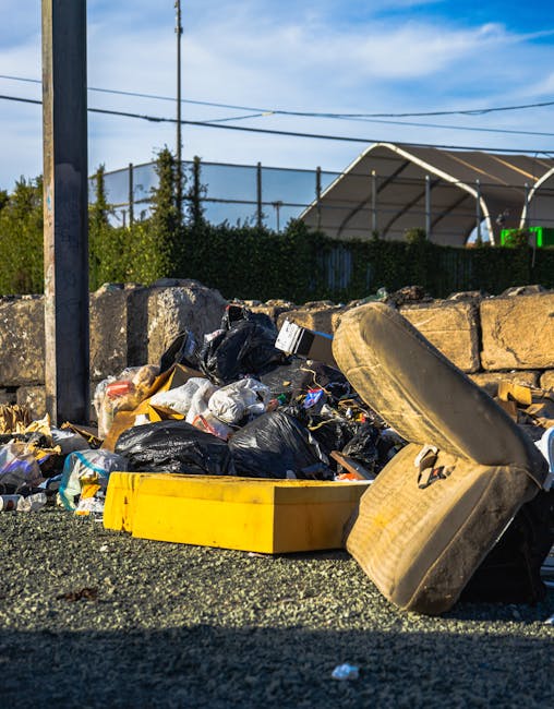 A close-up view of a collection of scattered rubbish on a gravel surface, including black and white plastic bin bags filled with waste, a worn beige upholstered chair with fabric torn and padding exposed, and a partially visible yellow plastic container or tray. The rubbish is situated near a low stone wall made of rough, uneven blocks, with some debris leaning against the wall. Behind the area, there is a background of green shrubbery and a tall metal fence, with a sports court or pavilion covered by a white canopy structure visible in the distance. Above, the sky is partly cloudy with hints of blue, and power lines are seen running across the scene. The setting suggests an outdoor space where waste has been improperly discarded, exemplifying the need for professional rubbish removal services such as those offered by Waste Disposal Catford, especially when multiple waste types and non-standard items are involved, indicating the importance of alternative waste handling solutions to avoid private and unauthorised dumping.