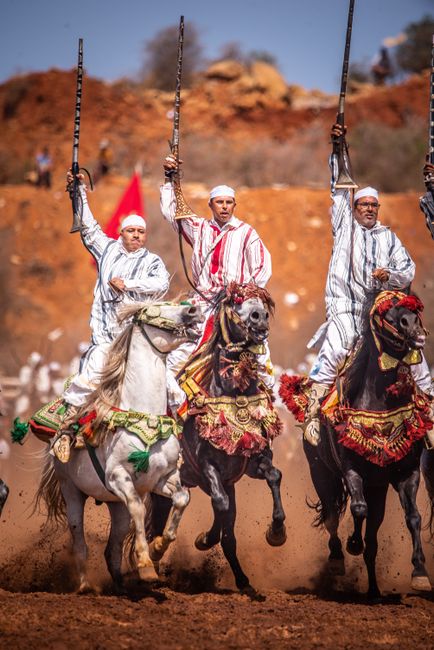 Three horse riders dressed in traditional striped white and red garments and white turbans are participating in a cultural event on a dusty, reddish-brown terrain. The riders are seated on elaborately decorated horses adorned with ornate, colorful saddlecloths and embroidered harnesses featuring floral and geometric patterns in red, green, and gold. Each rider is holding a long, slender spear upright in their right hand, with expressions of focus on their faces. The background reveals a landscape of reddish-brown rocky hills with sparse vegetation, under a bright blue sky. Dust kicked up by the horses' hooves rises into the air, enhancing the dynamic movement of the scene. The environment suggests an outdoor performance or festival, capturing a moment of traditional equestrian display associated with cultural or ceremonial celebrations, which may be linked to regions known for equestrian events and community gatherings, such as in parts of North Africa or the Middle East. This scene is unrelated to rubbish removal services, but visually demonstrates cultural practices involving horses and traditional attire, as seen in various forms of private or community-organized events in rural or semi-urban settings.