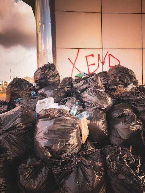 A close-up view of a collection of scattered rubbish on a gravel surface, including black and white plastic bin bags filled with waste, a worn beige upholstered chair with fabric torn and padding exposed, and a partially visible yellow plastic container or tray. The rubbish is situated near a low stone wall made of rough, uneven blocks, with some debris leaning against the wall. Behind the area, there is a background of green shrubbery and a tall metal fence, with a sports court or pavilion covered by a white canopy structure visible in the distance. Above, the sky is partly cloudy with hints of blue, and power lines are seen running across the scene. The setting suggests an outdoor space where waste has been improperly discarded, exemplifying the need for professional rubbish removal services such as those offered by Waste Disposal Catford, especially when multiple waste types and non-standard items are involved, indicating the importance of alternative waste handling solutions to avoid private and unauthorised dumping.