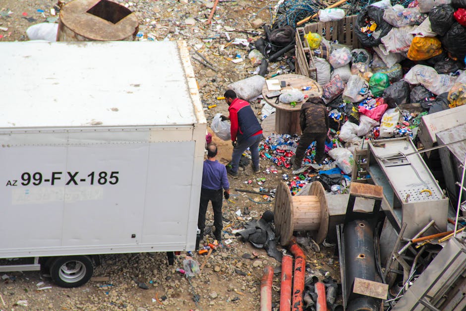 An outdoor scene showing a large white waste storage container with black alphanumeric markings, situated amidst a cluttered area filled with various types of rubbish. Surrounding the container, there are three individuals actively engaged in clearing the waste; one wearing a red and black jacket, another in dark clothing, and a third person with a pink-haired cap. The ground is covered in mixed debris, including plastic bottles, paper, and small discarded items, with scattered larger objects such as a wooden spool, metal pipes, and broken appliances. To the right, a pile of tightly packed garbage bags in black, white, and various bright colors is visible, along with loose trash and some wooden pallets. The scene appears to be part of an on-site waste clearance operation, reflecting an environment of manual rubbish removal often undertaken as an alternative to regular rubbish collection, highlighting the physical scale and mess associated with waste disposal activities. Waste Disposal Catford’s presence suggests professional intervention for complex waste handling. The background includes dirt, rocks, and debris, contributing to the chaotic and unmanaged appearance typical of large rubbish accumulation areas.